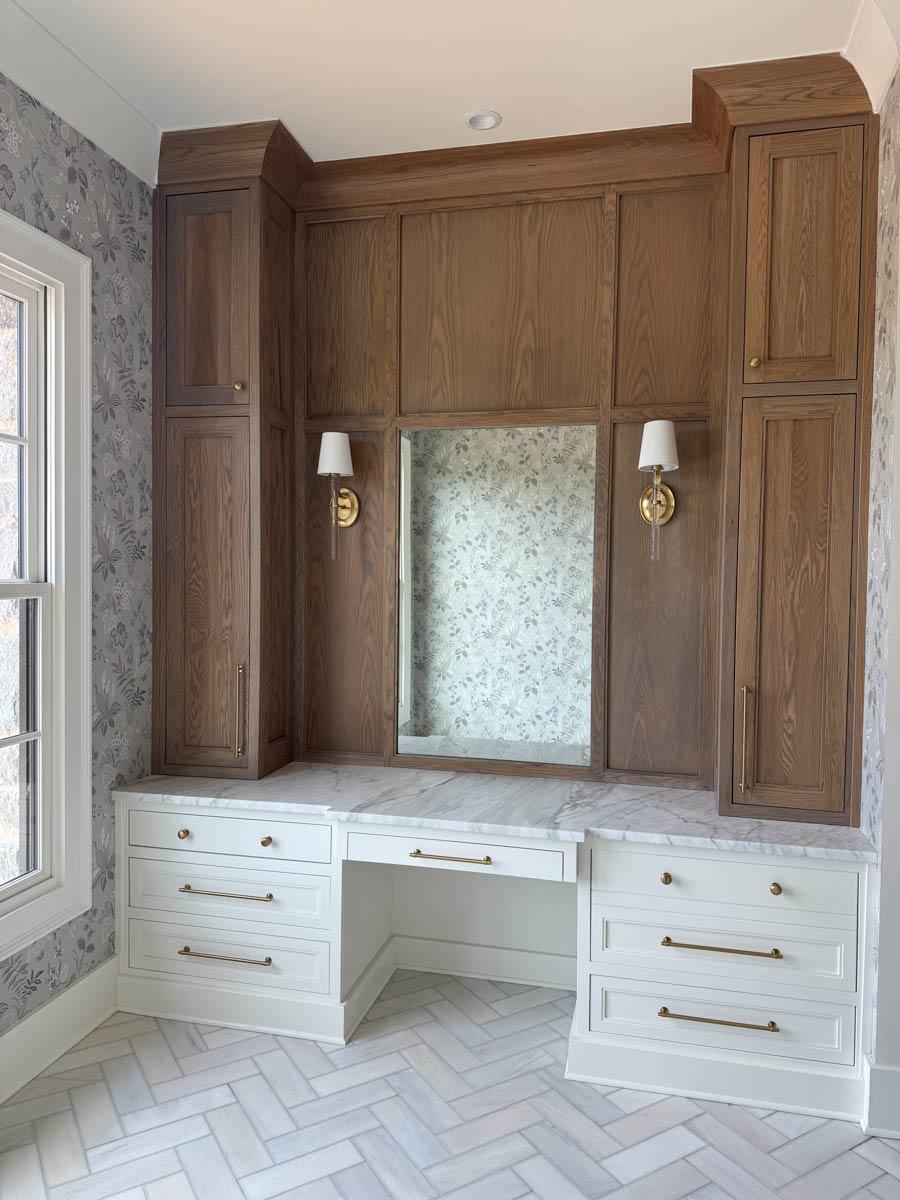 Built-in desk with white drawers, marble countertop, wood cabinetry, sconces, and floral wallpaper.