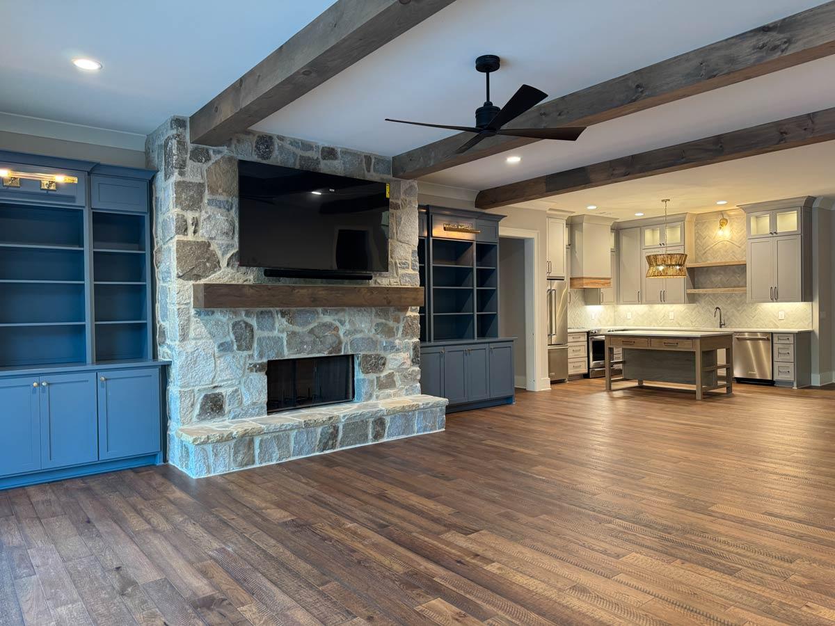 Great room with stone fireplace, built-in shelving, and open-concept kitchen with island and wood ceiling beams.