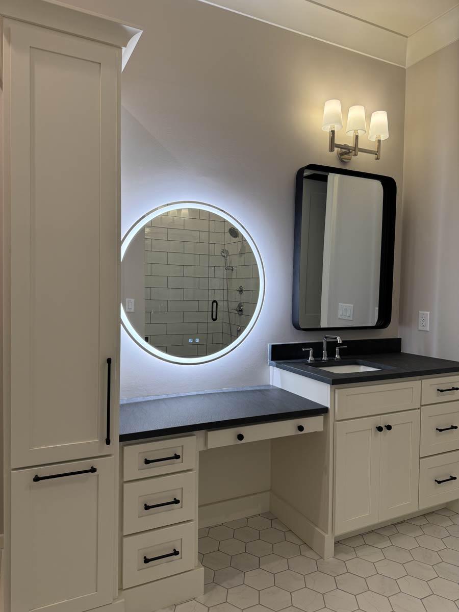 Bathroom vanity with a makeup counter, built-in storage, lighted circular mirror, and hexagonal floor tiles.