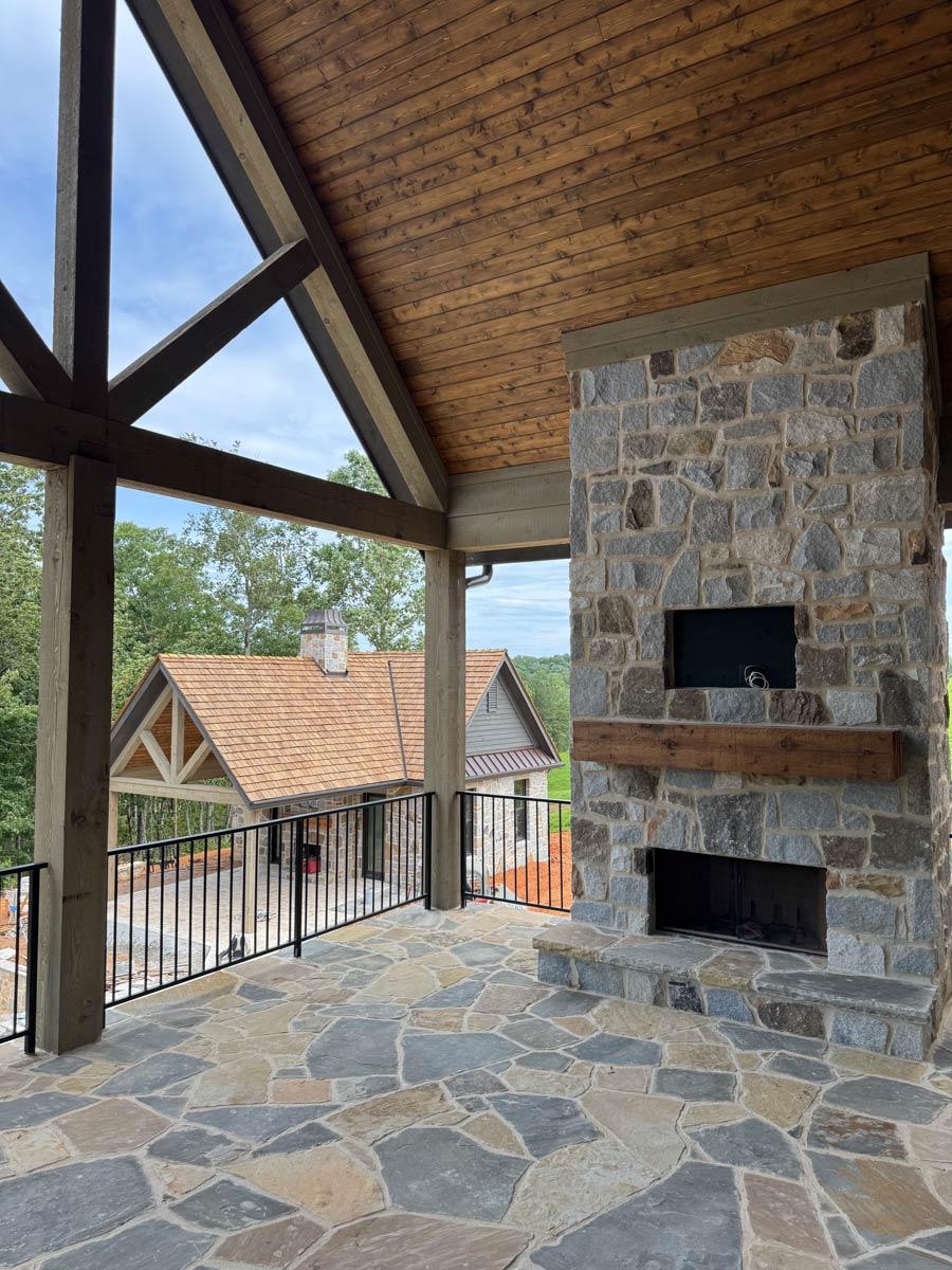 Covered porch with stone fireplace, vaulted timber ceiling, wood plank ceiling, and flagstone flooring. House exterior visible.