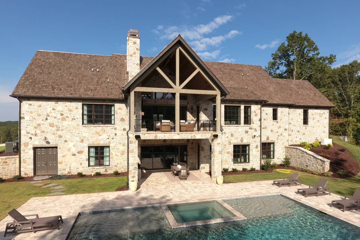 House plan exterior with stone facade, large gabled covered porch with exposed beams, and multiple window arrangements.