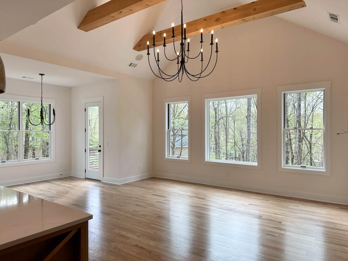 Interior view of open living area with vaulted ceiling, exposed beams, wood floors, large windows, and two chandeliers.