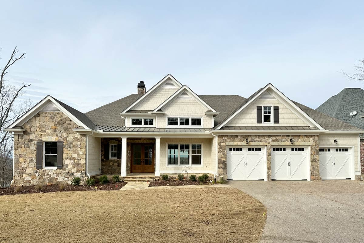 A large, newly built house with stone and cream siding, multiple gables, and white garage doors. The house is on a grassy, sloped lot.