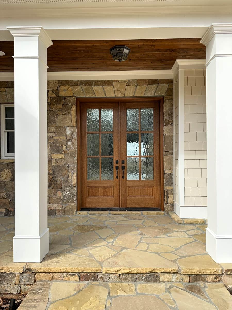 House plan exterior: Double wood doors with glass panes, stone and shingle siding, large porch with stone patio and white columns.