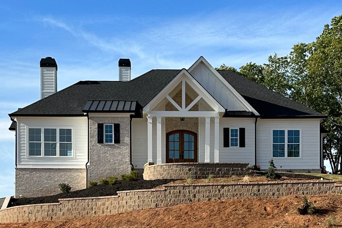 A newly constructed white house with a black roof and stone accents. The house has a gabled entry with a wooden door and sits on a tiered landscape.