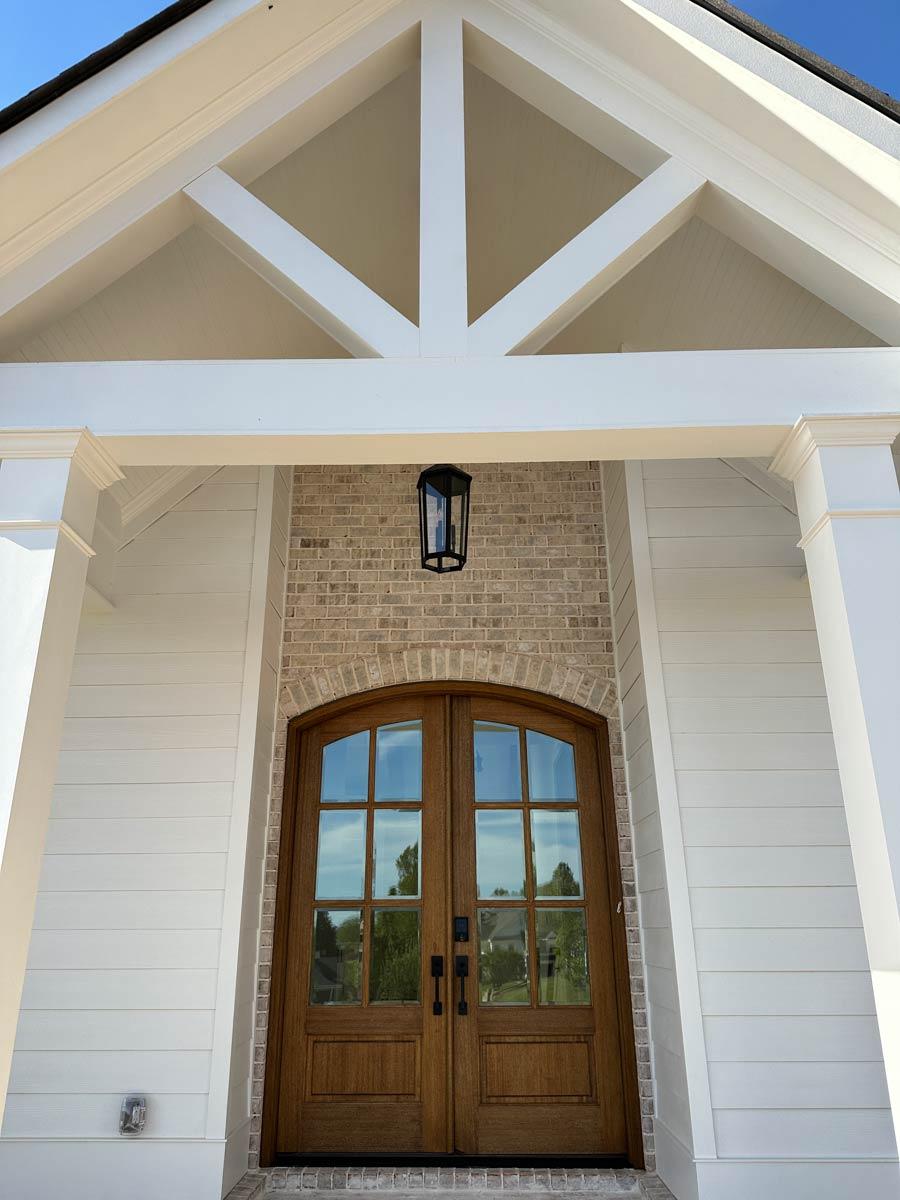Exterior house plan: Double wooden doors with arched glass panes, brick surround, white siding, and exposed rafter tails.