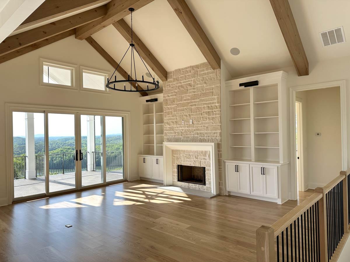 Great room interior with vaulted ceiling, exposed beams, stone fireplace, built-in bookshelves, and large sliding glass doors.
