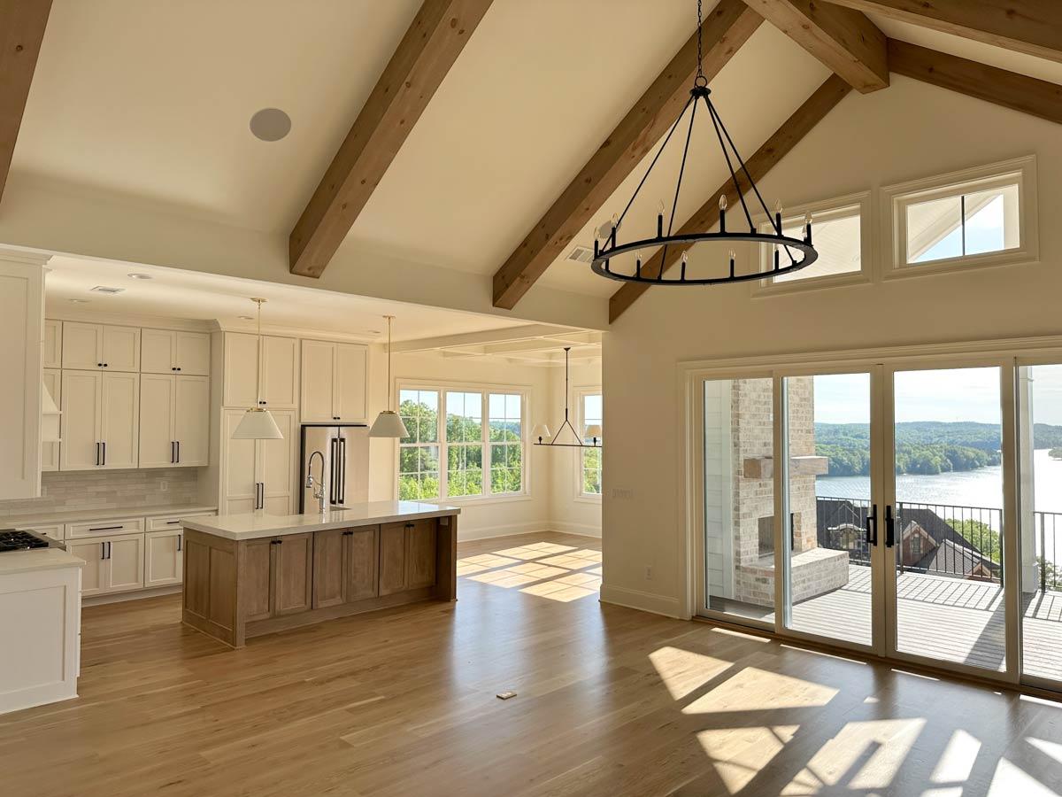 Open concept living space with vaulted beamed ceiling, kitchen island, fireplace, and large windows overlooking a lake.