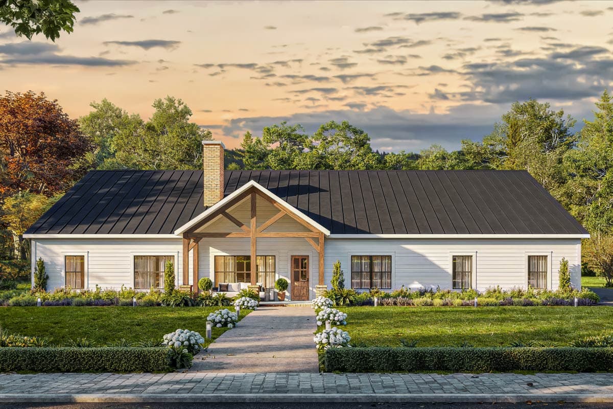 A white farmhouse with a dark roof and a porch with exposed wooden beams, set against a backdrop of trees and a cloudy sunset.