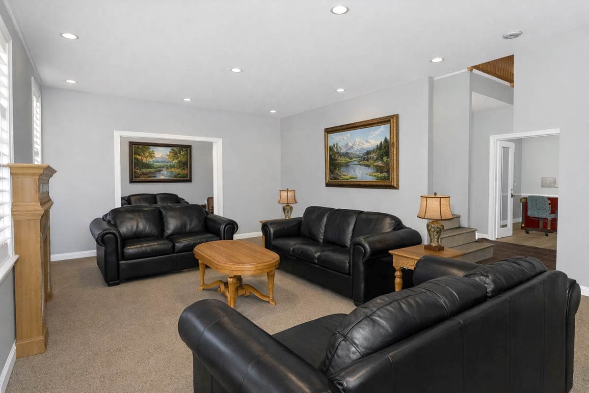 Living room with black leather sofas, wooden coffee table, fireplace, and landscape paintings. Doorway reveals a separate room with desk.
