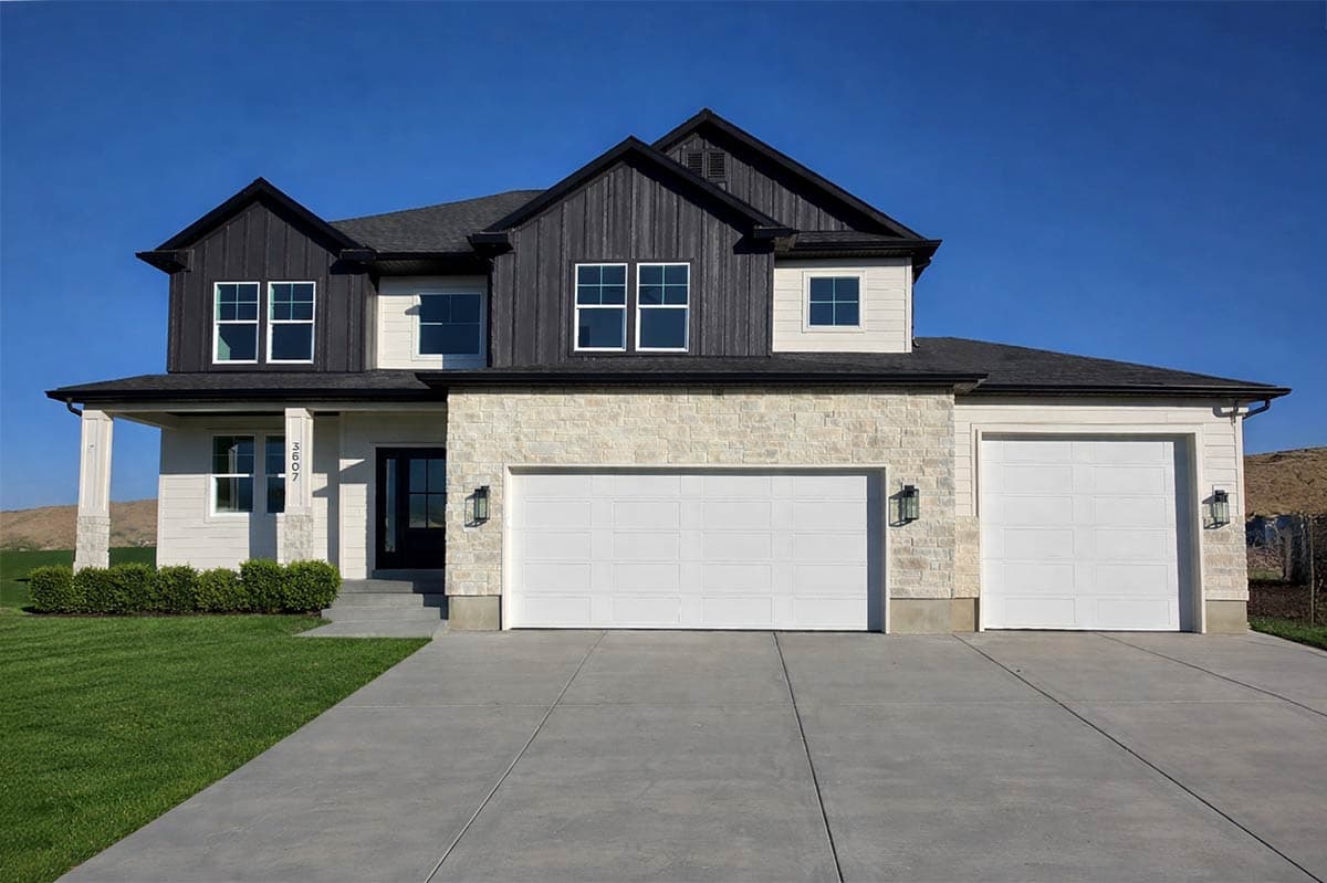 A modern two-story house with a contrasting black and white facade. Features include a stone-brick base, two garage doors, and a lush green lawn.