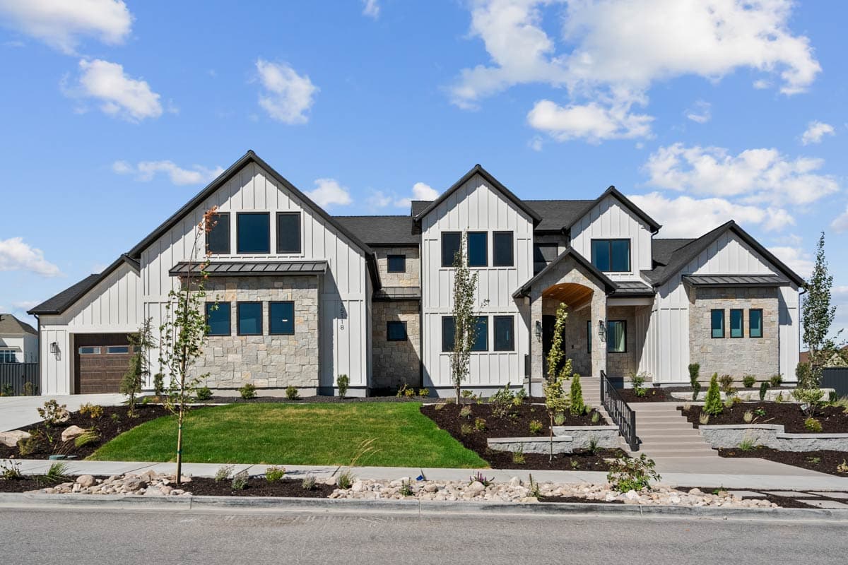 A large, modern house with a white exterior, black roof, and stone accents. It has multiple gables and sits on a well-manicured lawn with a blue sky.