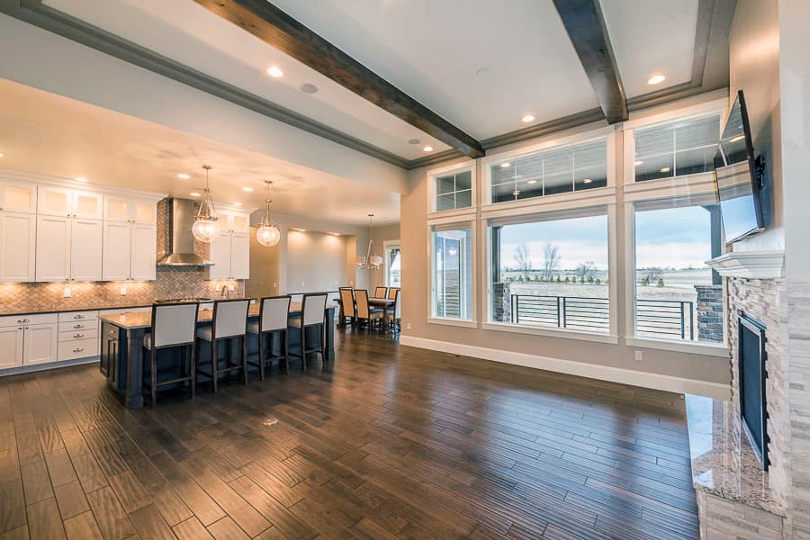 Open concept living area with kitchen island, large windows, stone fireplace, and exposed wood beams.