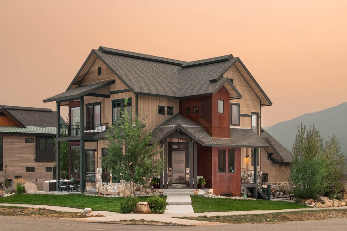 A modern two-story home with a mix of wood and rusted metal siding. The home features a dark roof, balcony, and sits on a well-manicured lawn.