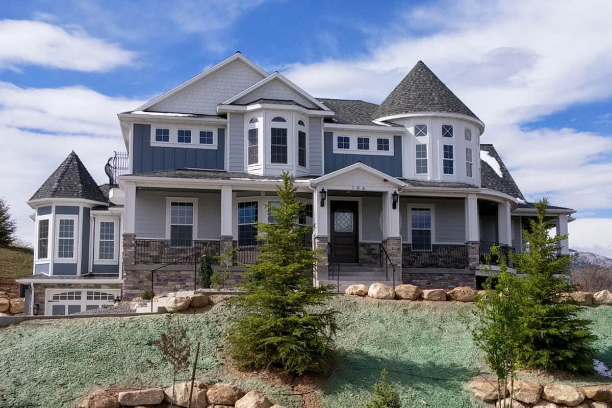 A large, two-story house with blue siding, multiple turrets and windows, a front porch, and a well-manicured lawn under a blue sky.