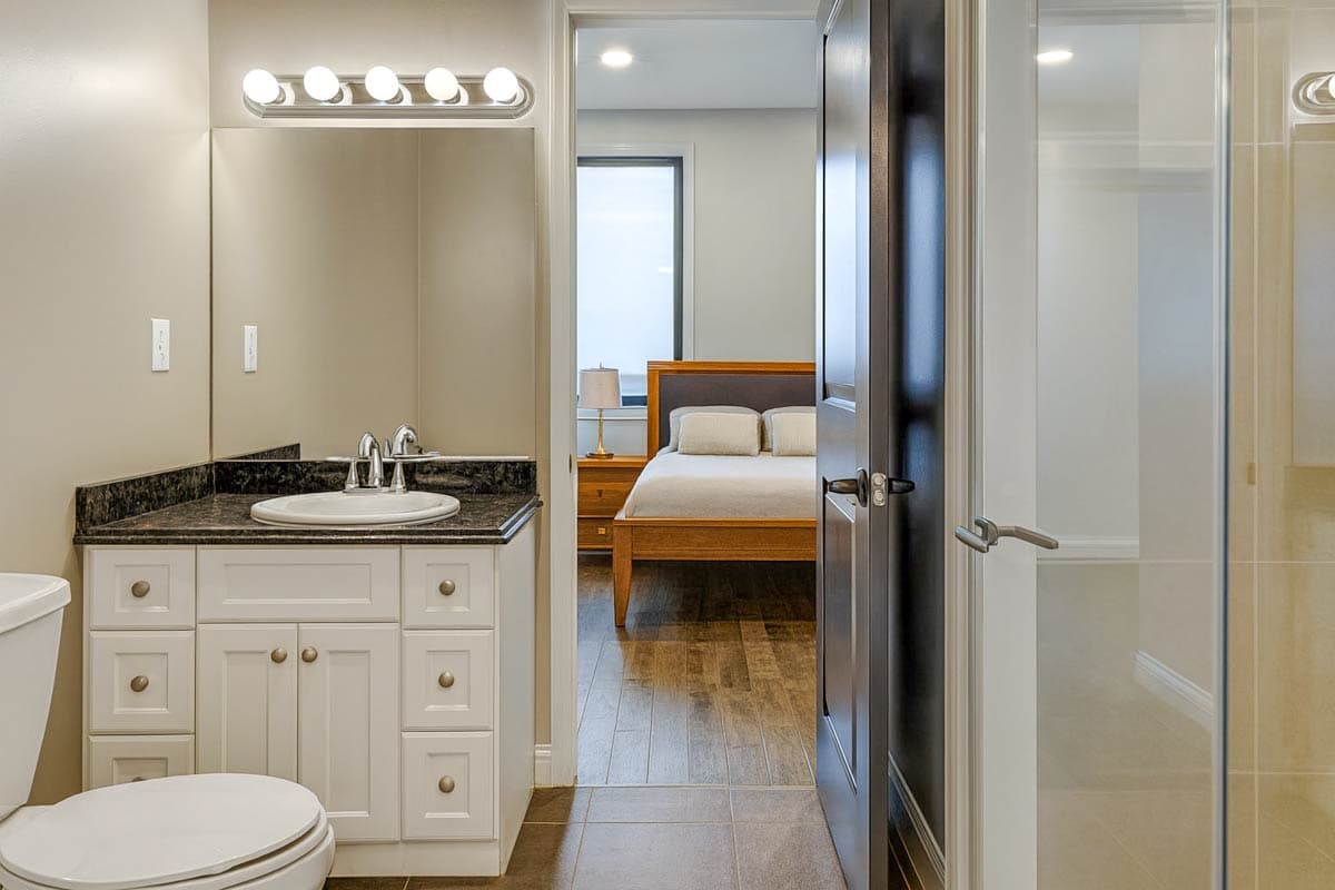 View into bedroom from bathroom with vanity, toilet, and glass-enclosed shower.