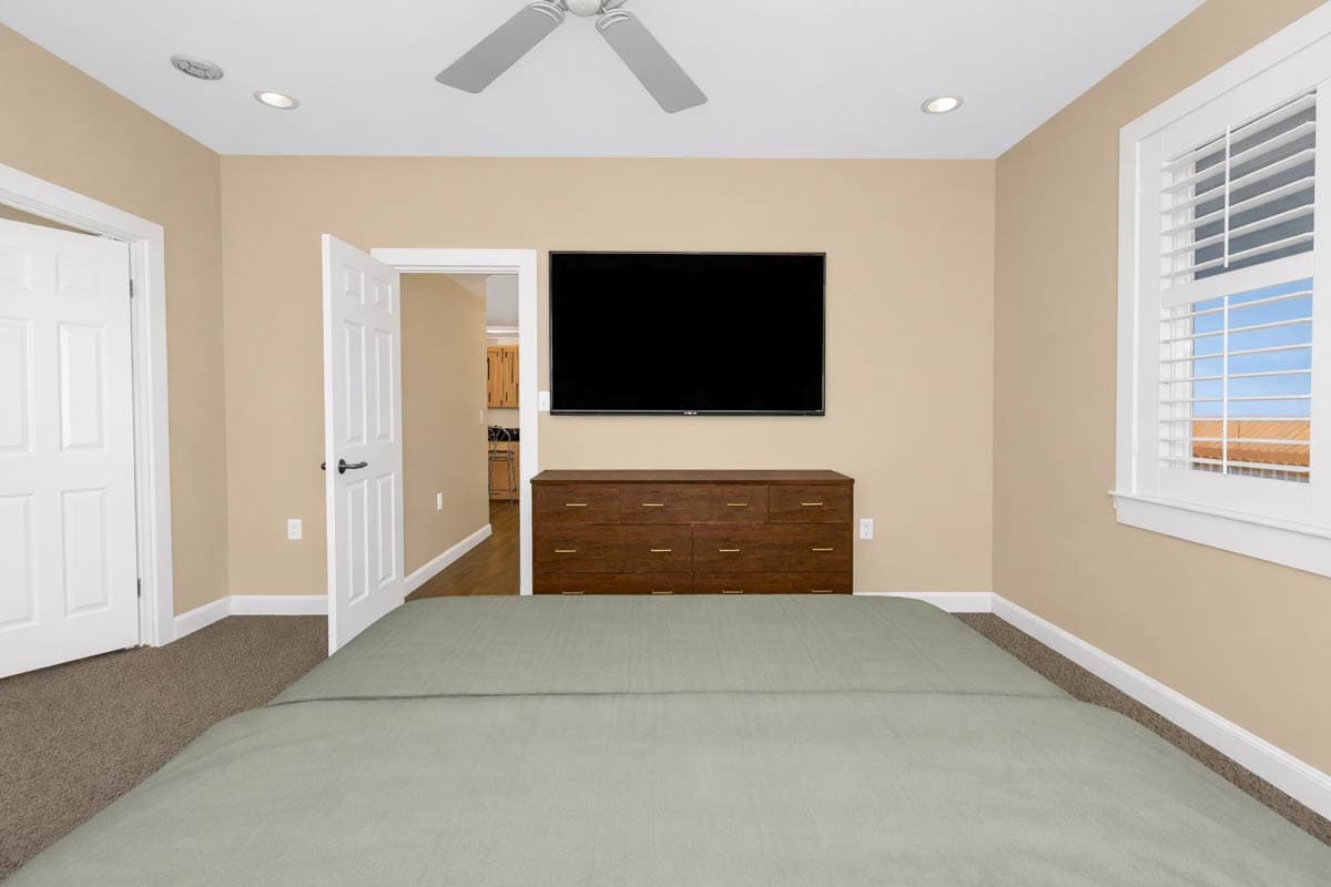 Bedroom interior with ceiling fan, mounted TV, dresser, and plantation shutters on window. Open door reveals adjacent room.