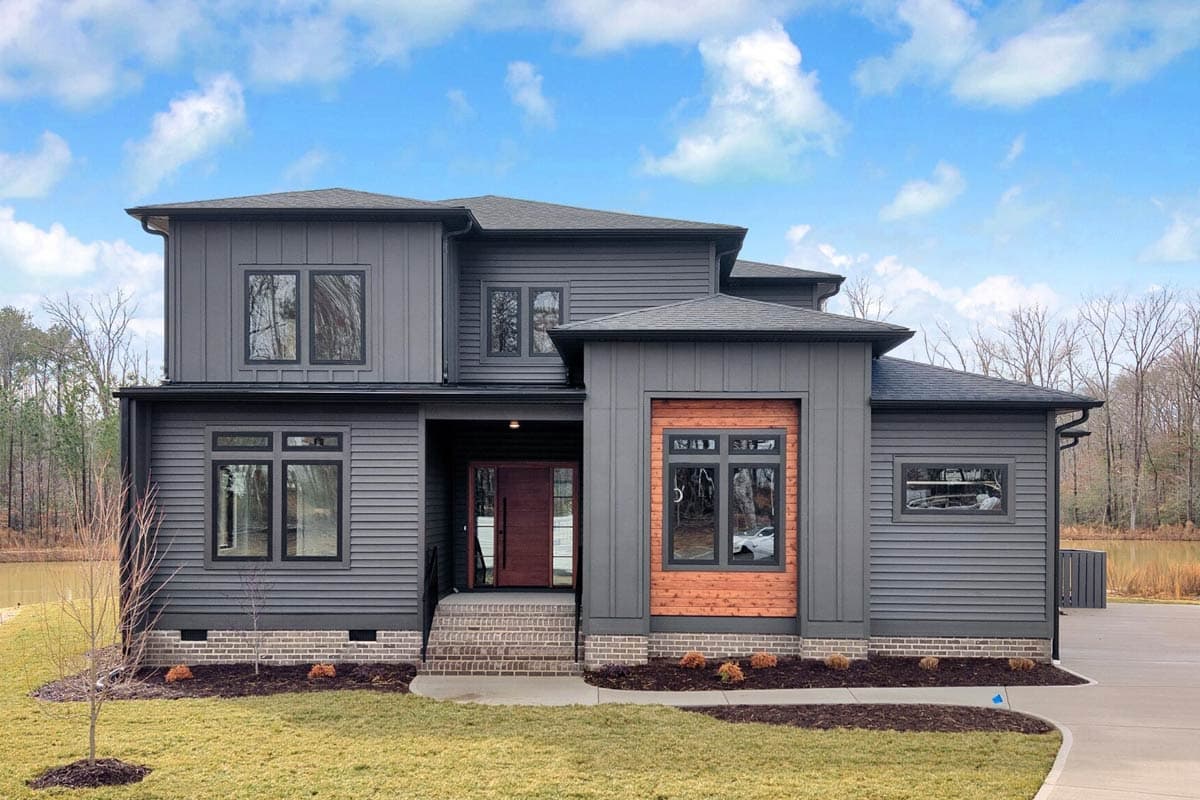 Modern two-story home with gray siding, black-framed windows, and a dark wood front door. Brick accents and a small yard with a cloudy blue sky.