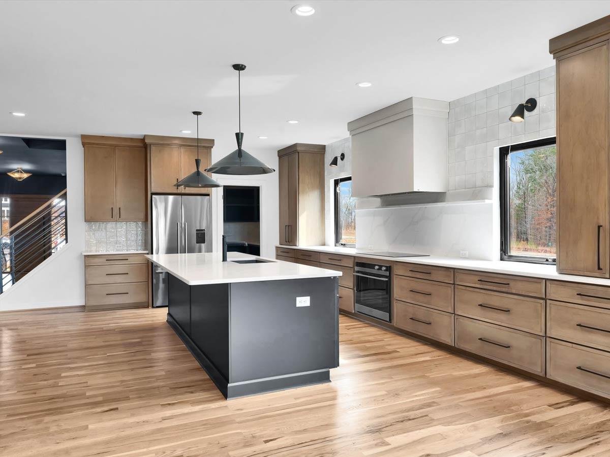 Modern kitchen with island, stainless steel appliances, wood cabinetry, and large windows. View of staircase to the left.