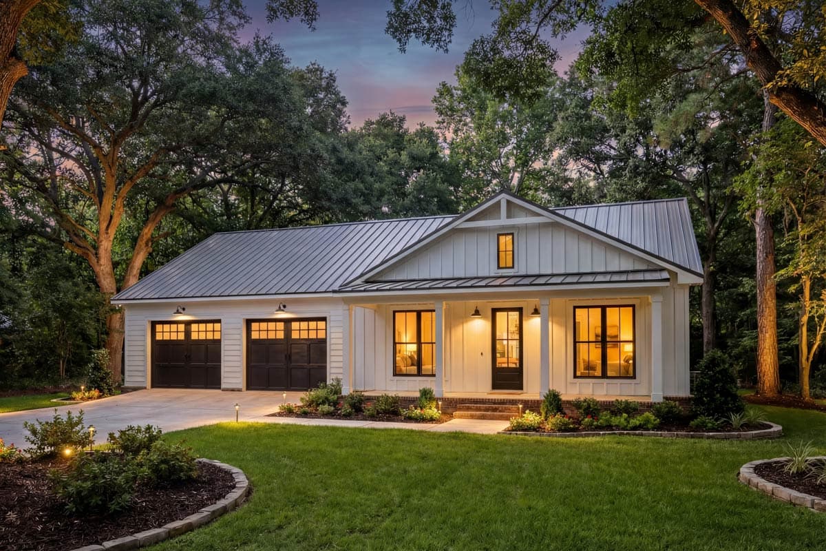 White farmhouse-style home with a dark metal roof. Lit windows and garage doors with mature trees in the background set in a green lawn.