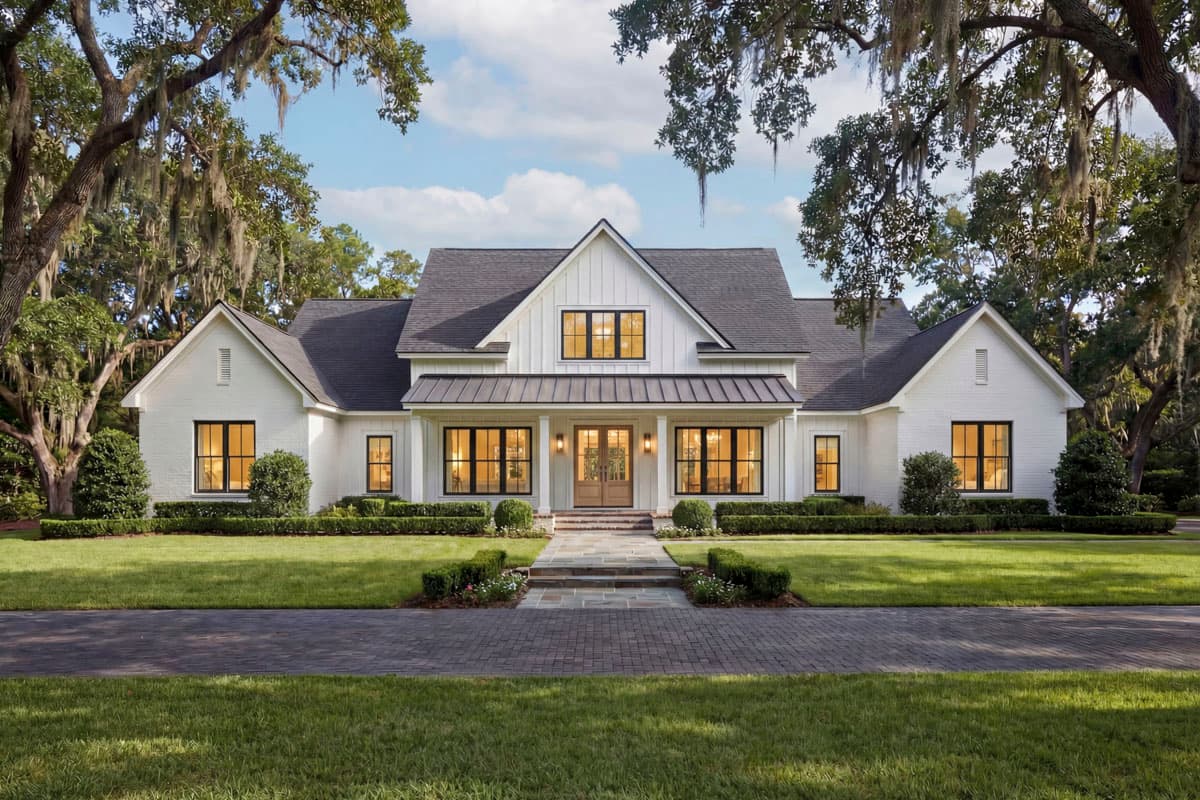 A white farmhouse with a dark roof and black window frames is set amidst lush green landscaping and mature oak trees, creating a serene environment.