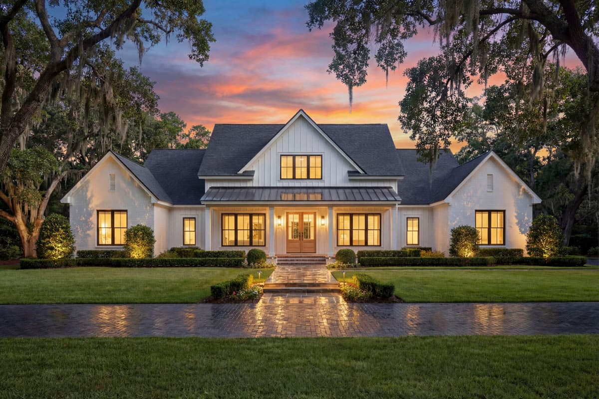 A white farmhouse with black trim glows at dusk, its windows lit against a colorful sunset. Spanish moss hangs from surrounding trees.