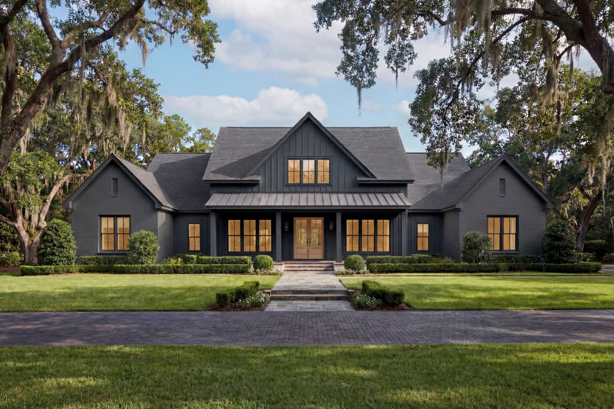 A modern, dark grey house with symmetrical design is framed by lush trees and a well-manicured lawn. A brick path leads to the front door.