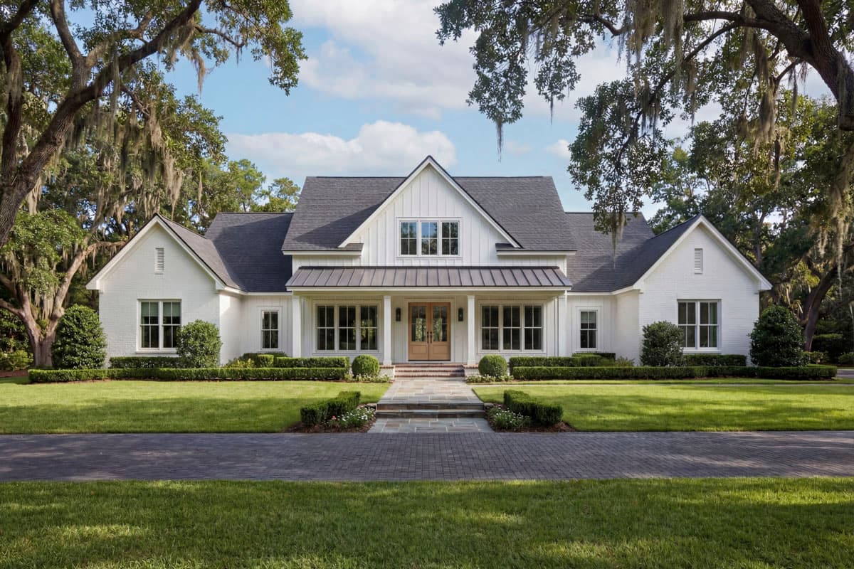 A white, two-story house with a dark gray roof is framed by large oak trees draped with Spanish moss and a manicured lawn.