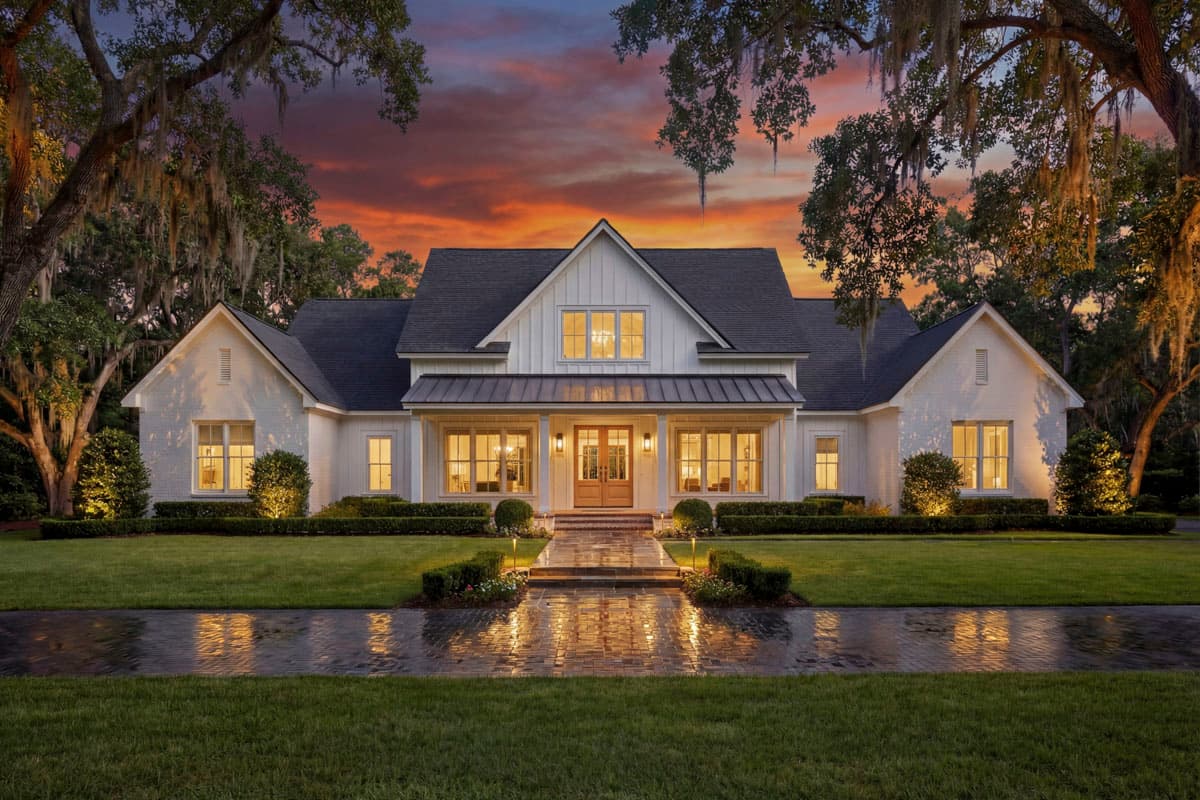 A white farmhouse with a dark roof is lit up at dusk. A paved walkway leads to the front door, lined by manicured bushes and green grass. Trees frame the house.