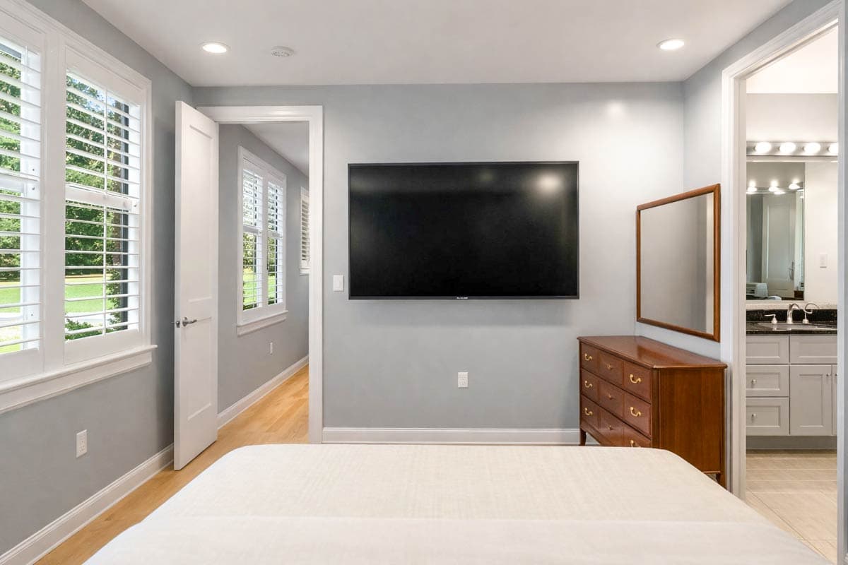 Bedroom with wall-mounted TV, dresser, and open doorway to bathroom. White plantation shutters on windows.