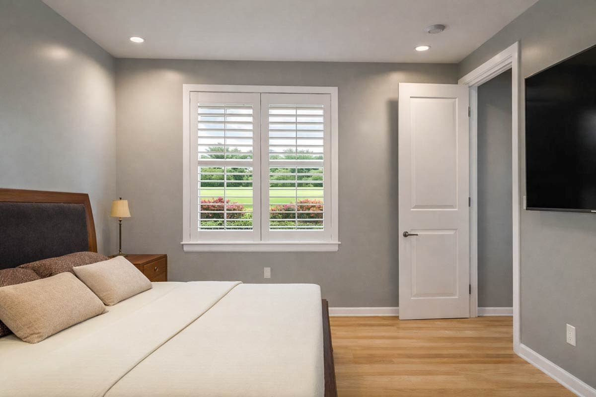 Interior view of bedroom with plantation shutters on window, wood floor, and open doorway.