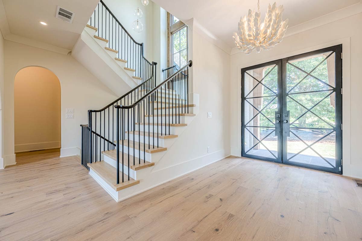 Interior view of house foyer with a curved staircase, black railing, arched doorway, and double glass doors with metal grilles.