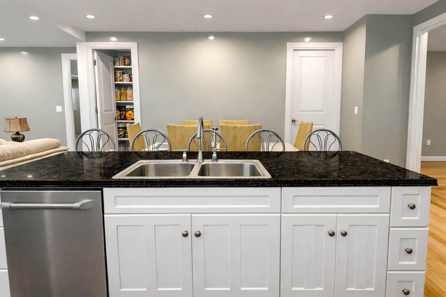 Kitchen island with dual sink, granite countertop, dishwasher, and white shaker cabinets. Adjacent dining area with chairs.