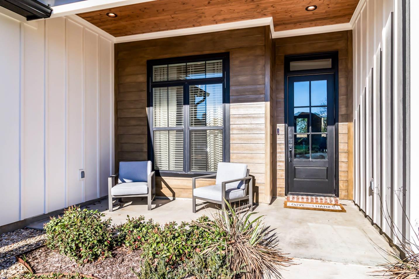 Modern Farmhouse house plan exterior with wood siding, covered porch, black door, and two outdoor chairs.