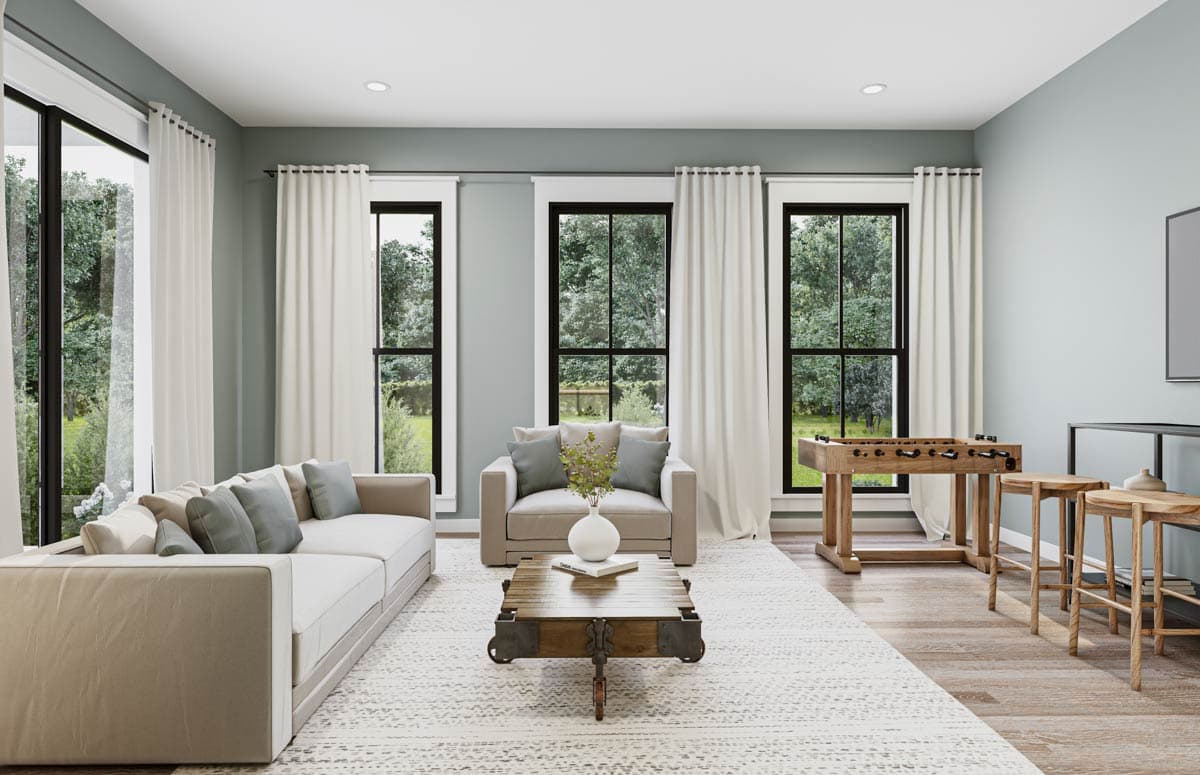 Living room with large windows, sofa, and a coffee table on a patterned rug. Includes foosball table and small side tables.