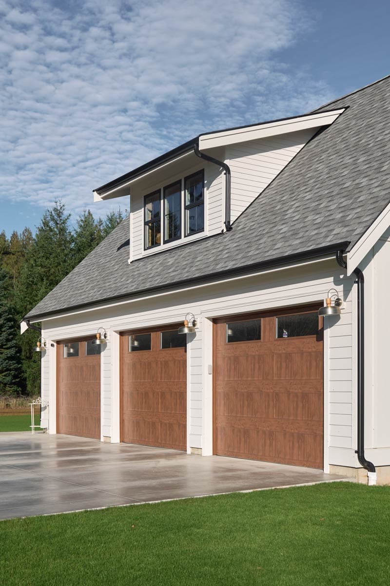 House plan exterior: Three wood-grain garage doors with glass transoms, dormer with triple windows, white siding, and gray shingle roof.
