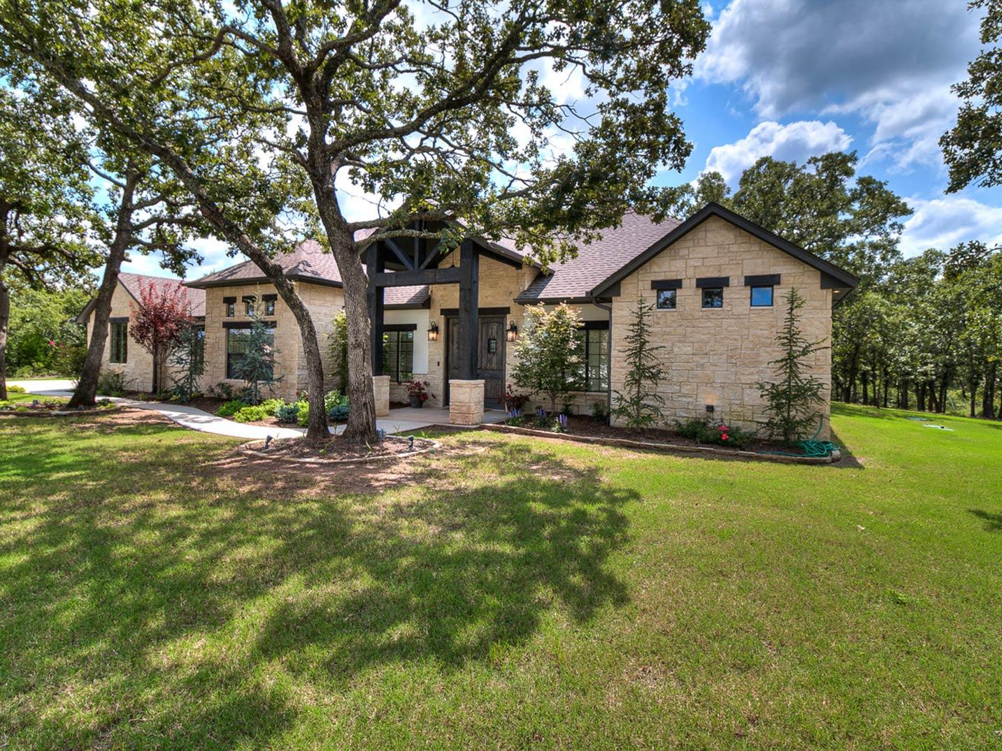 House plan exterior: Single-story stone home with dark wood-framed entrance, gable roof, and large trees.