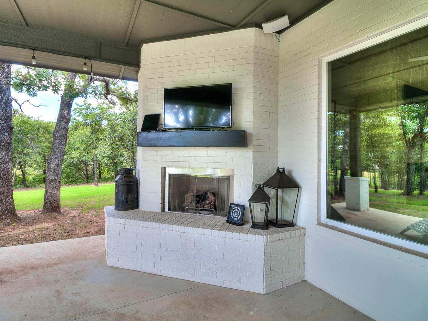 Covered patio with brick fireplace, TV, wood mantle, and large window overlooking trees and lawn.