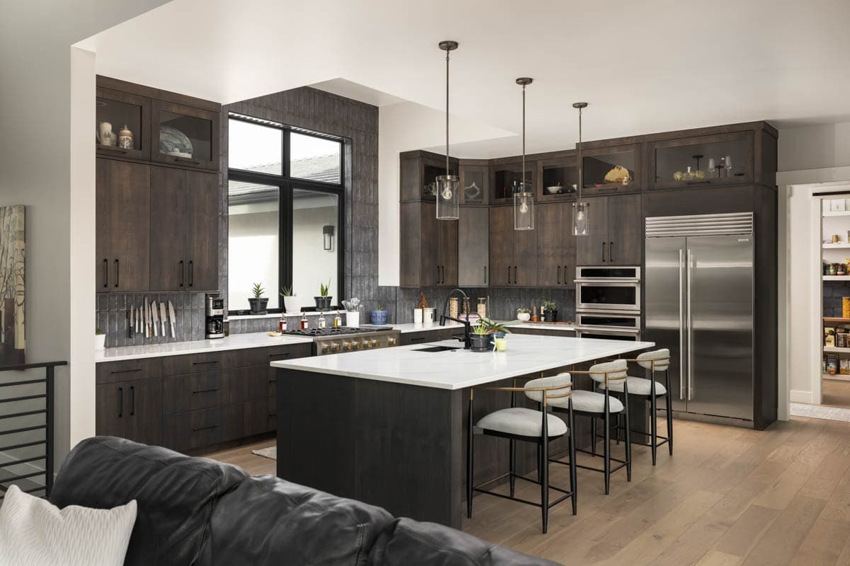 Modern kitchen with dark wood cabinetry, white quartz island, stainless steel appliances, and a large window.