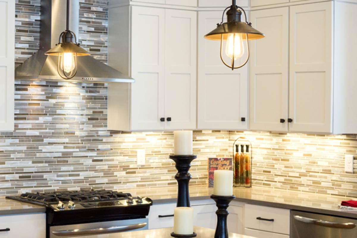 Kitchen interior with white shaker cabinets, mosaic tile backsplash, stainless steel range hood, and pendant lights.
