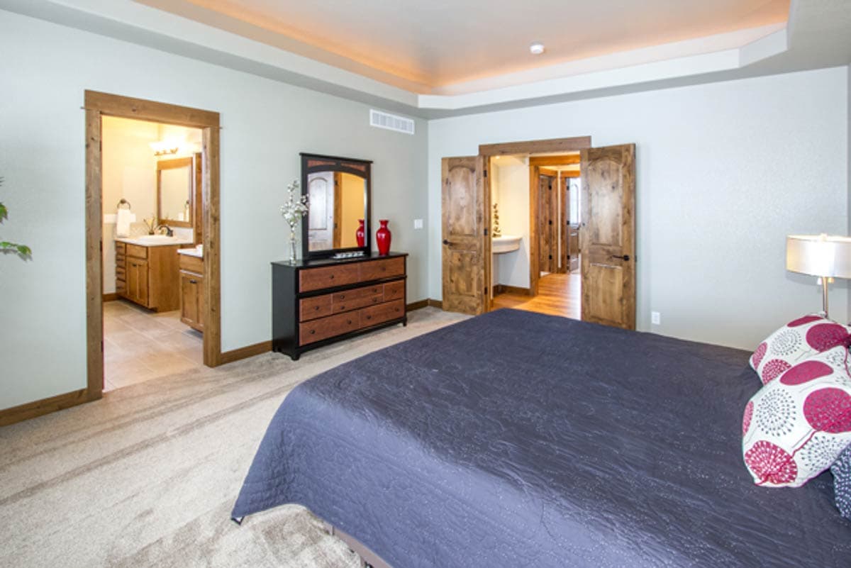 Bedroom interior with ensuite bathroom access through wooden double doors, dresser, and neutral carpeting.