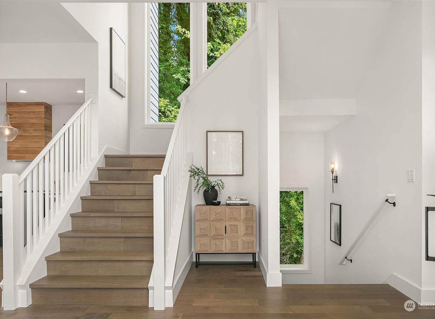 Interior view of a modern entryway with a wood staircase, white banisters, tall windows overlooking trees, and a geometric credenza.