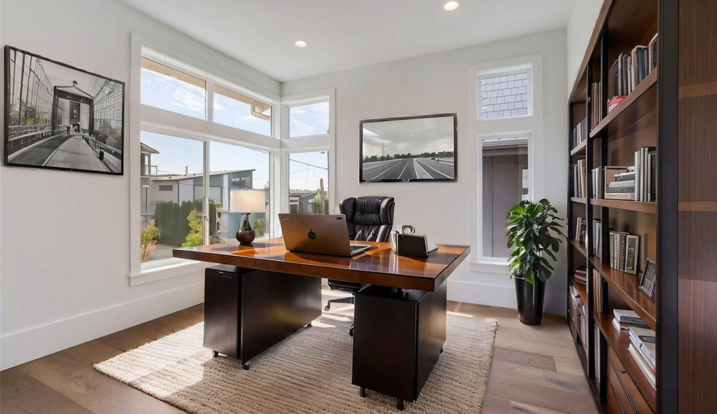 Home office with wooden desk, leather chair, floor-to-ceiling bookshelf, and large corner windows.