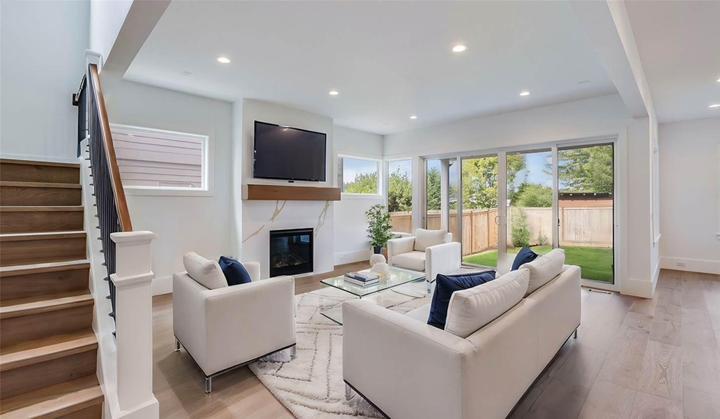 Modern living room with white sofas, marble fireplace, wood staircase, and sliding glass doors to backyard.