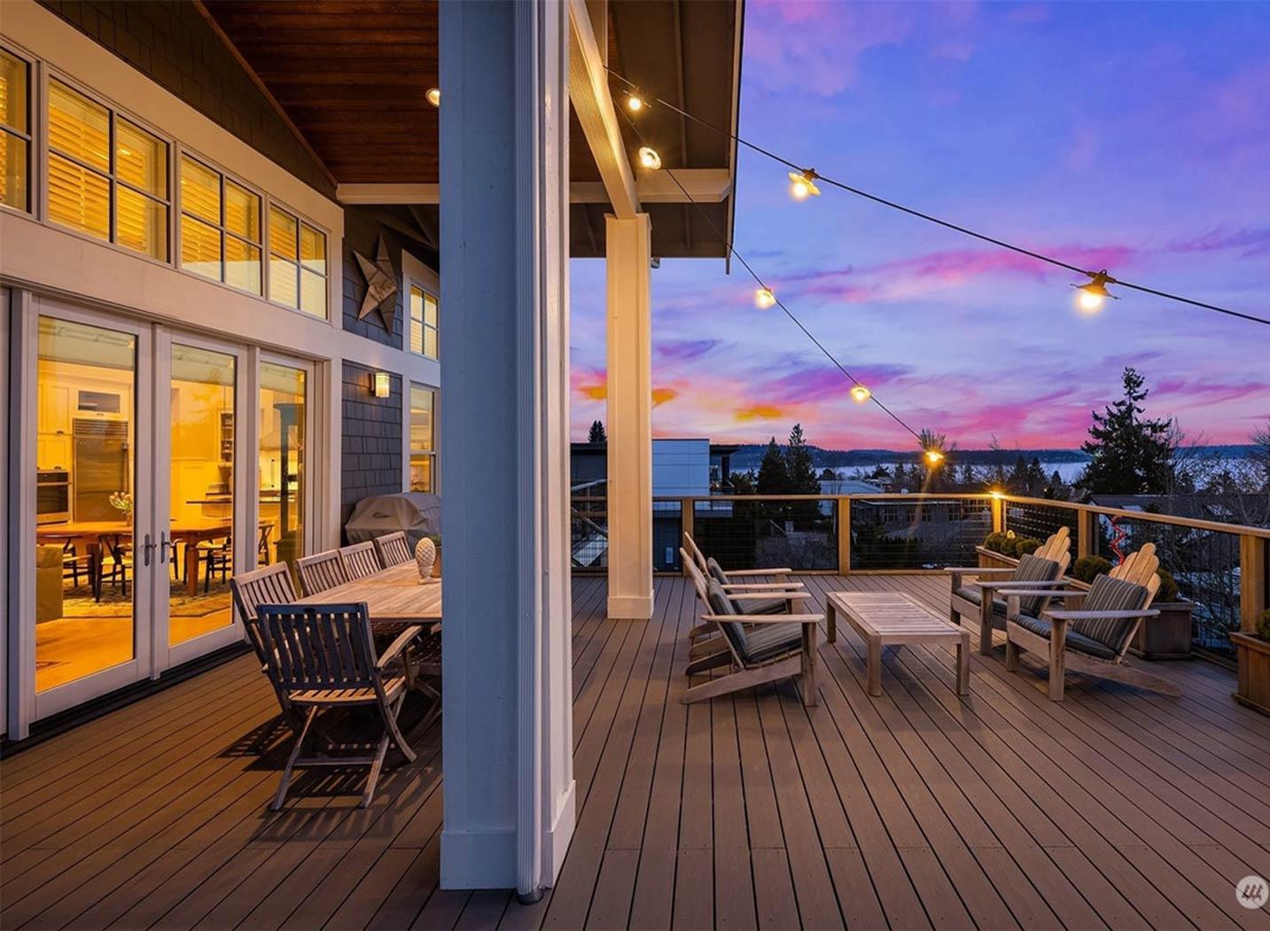 Covered deck with dining and seating areas, string lights, and expansive water views at sunset.