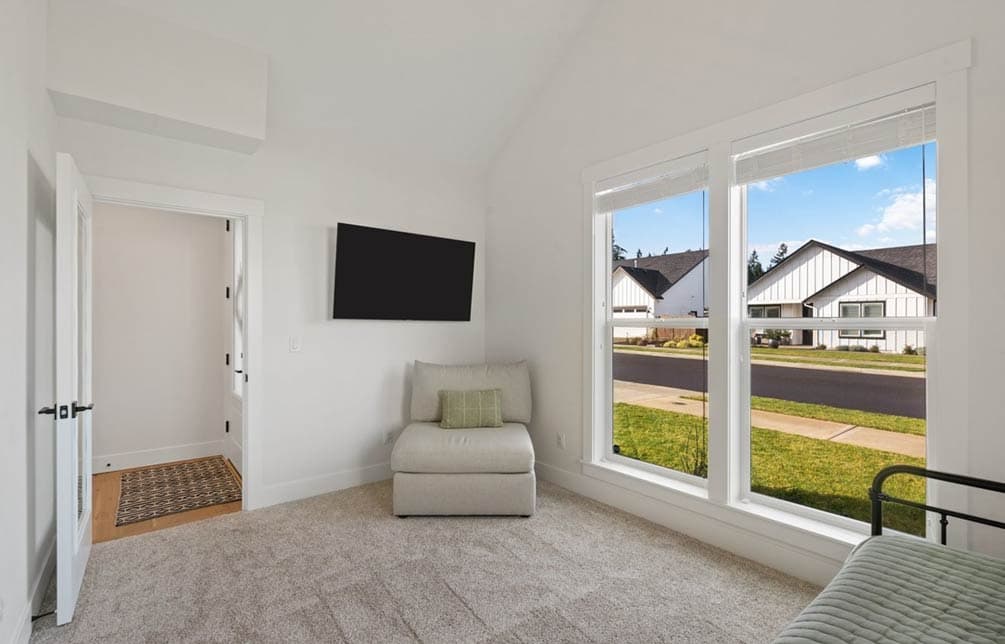 Interior view of a room with a wall-mounted television, armchair, and large windows showing houses.