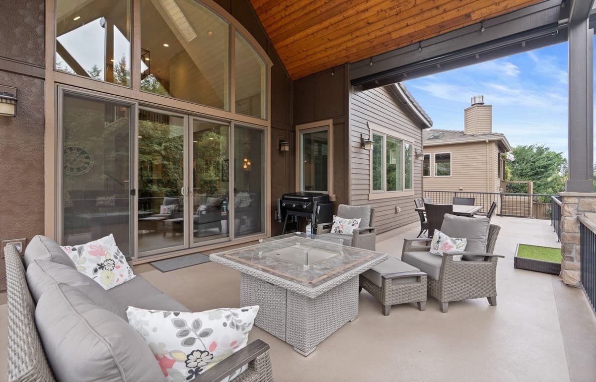 Covered outdoor living space with seating, fire pit, grill, and dining area; adjacent to house with large windows and wood ceiling.