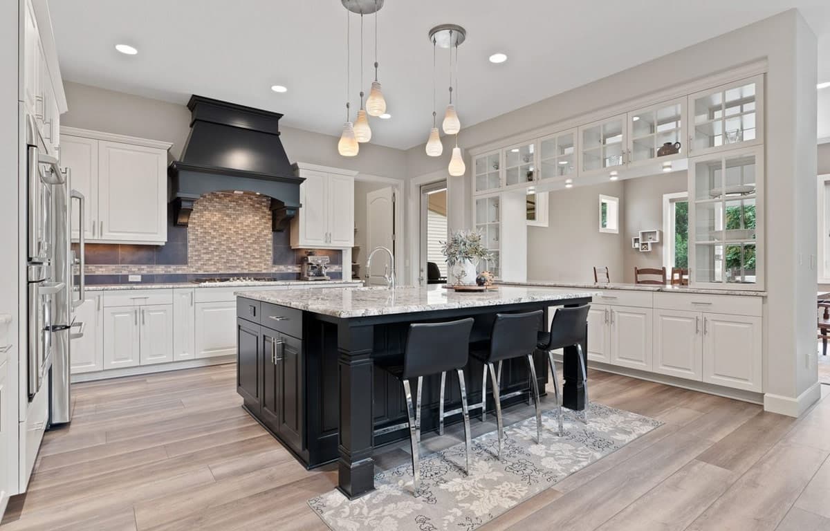 Kitchen with island, white cabinets, dark range hood with mosaic tile backsplash, and glass-front upper cabinets.