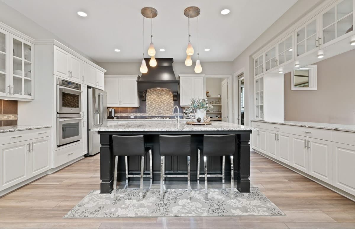 Kitchen with a large island, black bar stools, white cabinets, double ovens, and a dark range hood with tile backsplash.