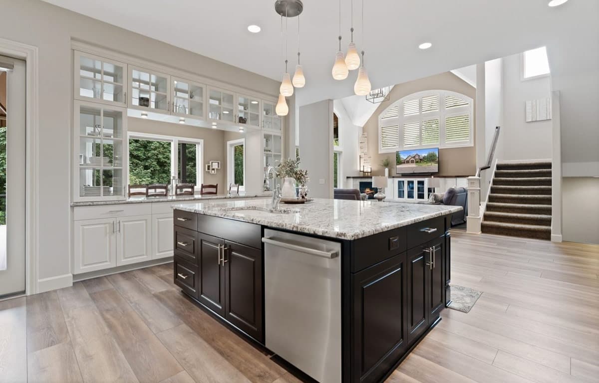 Interior view of a modern kitchen with dark island, granite countertops, white cabinetry, and open-concept living area with fireplace and stairs.